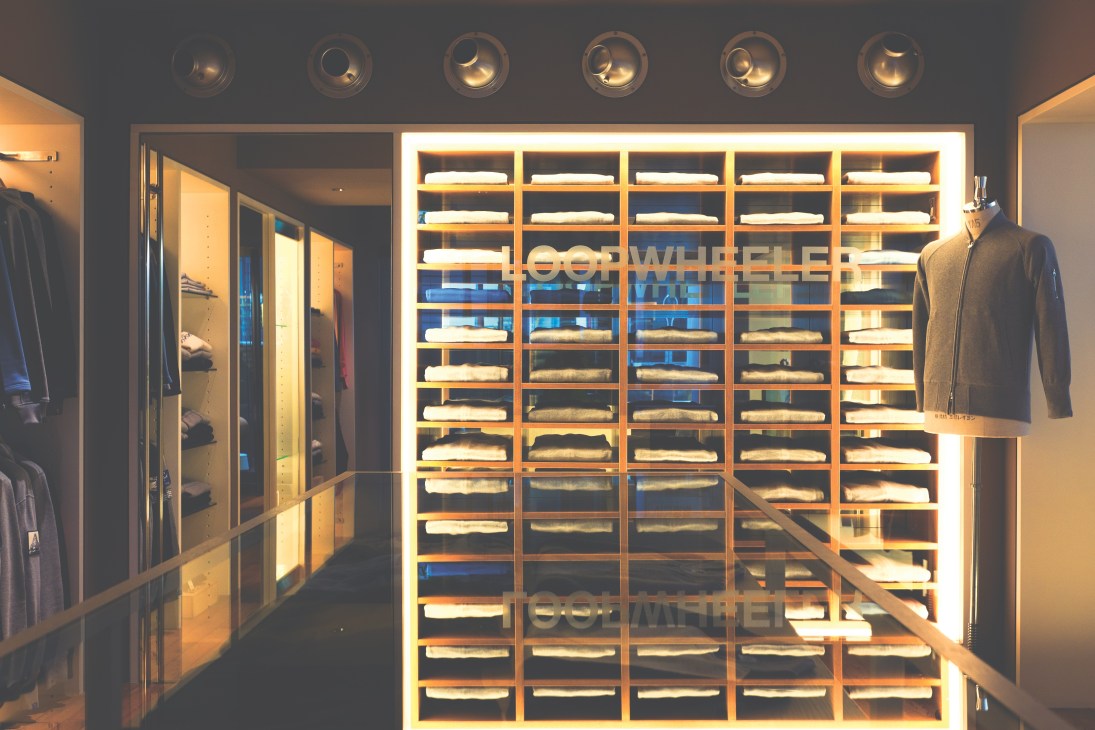 Loopwheeler's flagship Tokyo shop interior with illuminated wooden shelving displaying neatly stacked sweatshirts and cardigans, with garments on mannequins.