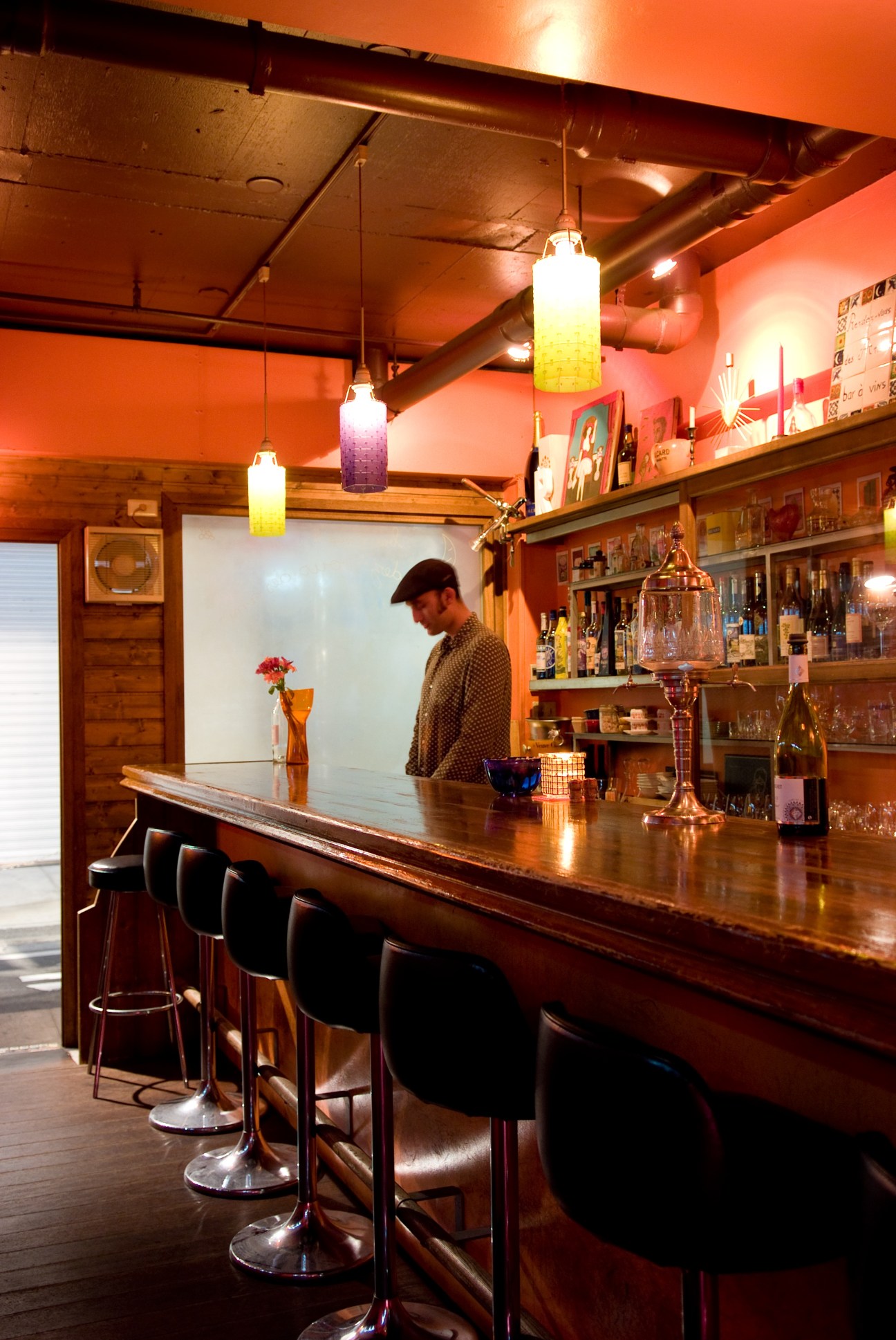 Mathias Incandela stands behind the wooden bar counter of Rendezvous des Amis, a stylish bar with warm orange walls, hanging colored glass pendant lights, and shelves of bottles.