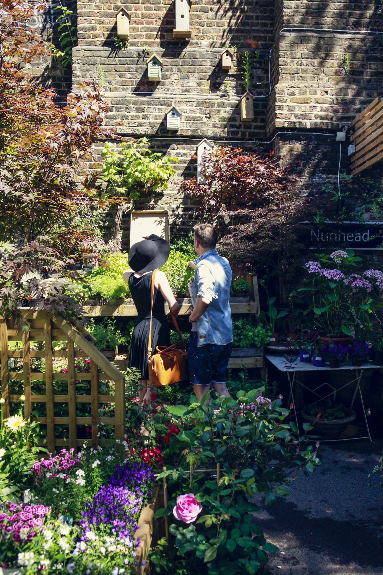 Two people browse plants at The Nunhead Gardener garden centre in London, surrounded by colorful flowers and greenery against a brick wall.