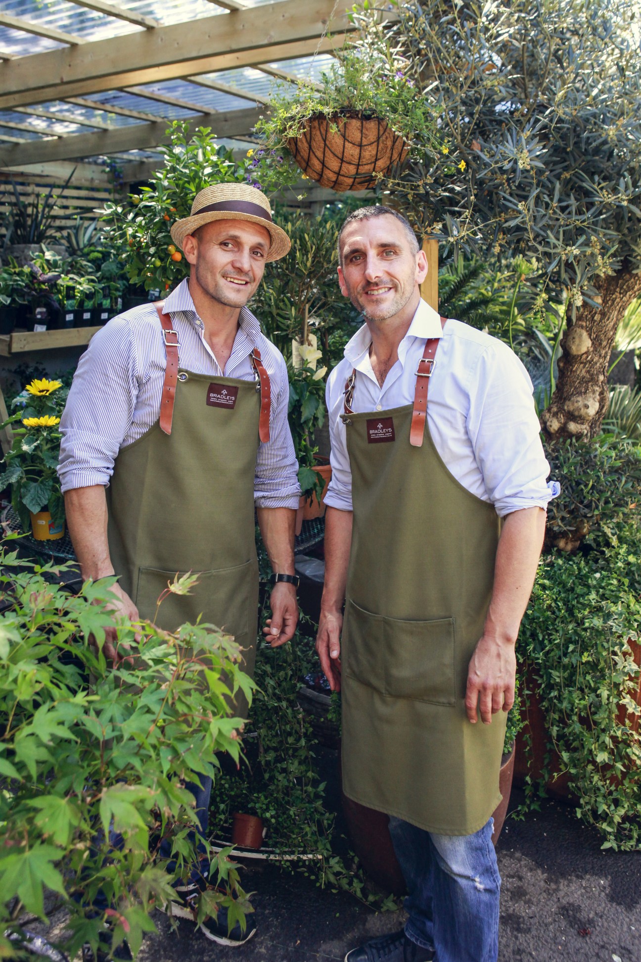 Two men wearing olive-green aprons stand among plants at The Nunhead Gardener garden centre in London.
