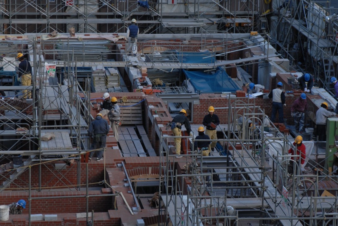 Construction workers in hard hats assembling red brick walls and steel scaffolding on a Marunouchi building site.