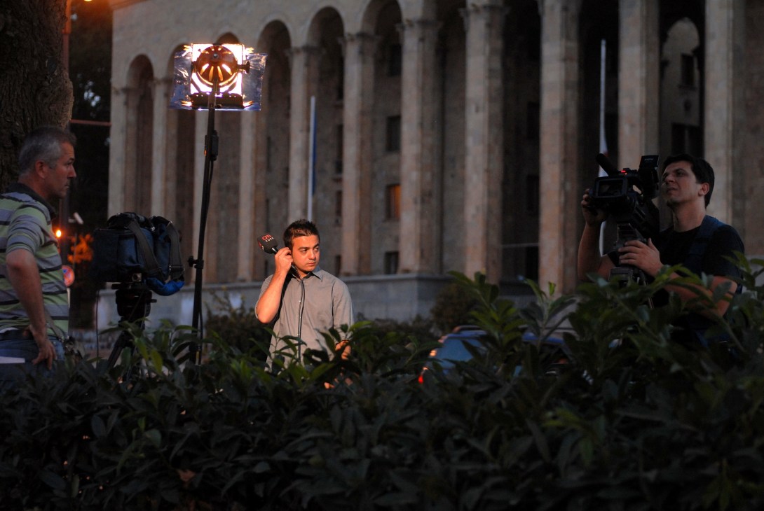 A Turkish CNN anchor reporting live at night in Tbilisi with camera crew and lighting equipment.