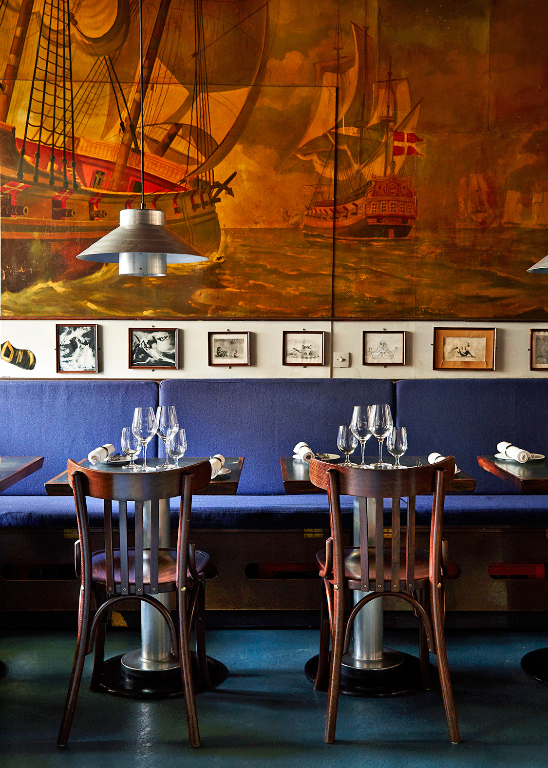 Restaurant dining room with wooden chairs, blue banquette seating, place settings, and nautical mural artwork.