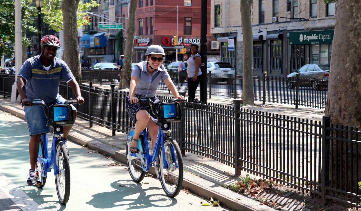Two people riding blue Citi Bike bicycles on a tree-lined New York City street.