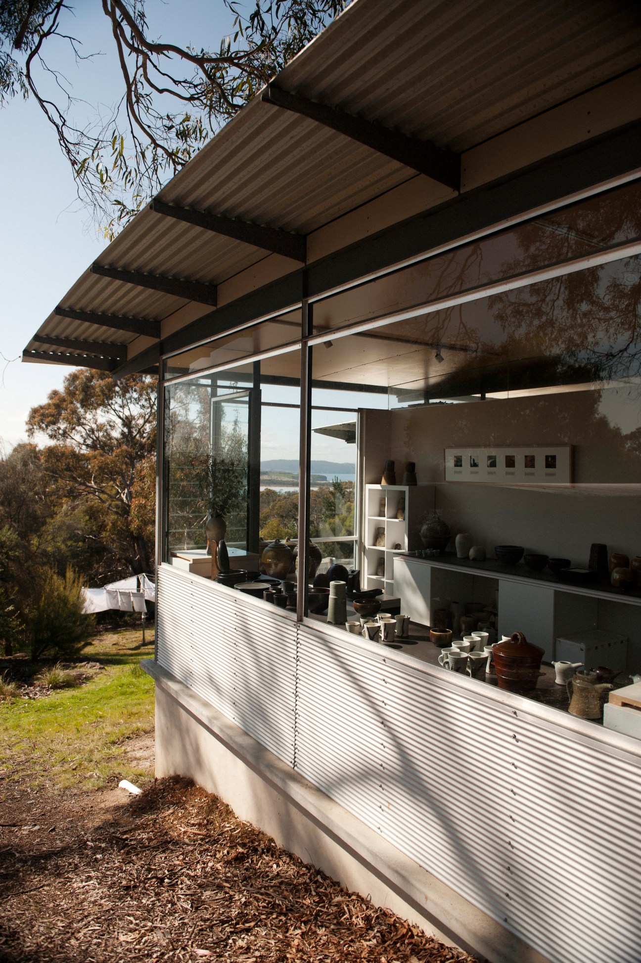 Modern pottery studio with corrugated metal roof, glass doors and shelving displaying ceramic work overlooking a lagoon.