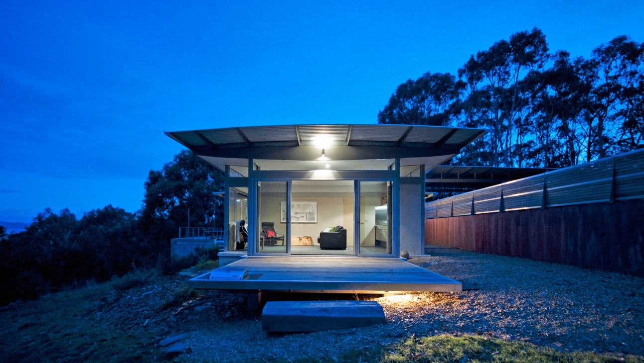 Modern minimalist guesthouse with glass front, lit interior, and wooden deck at dusk surrounded by eucalyptus trees.