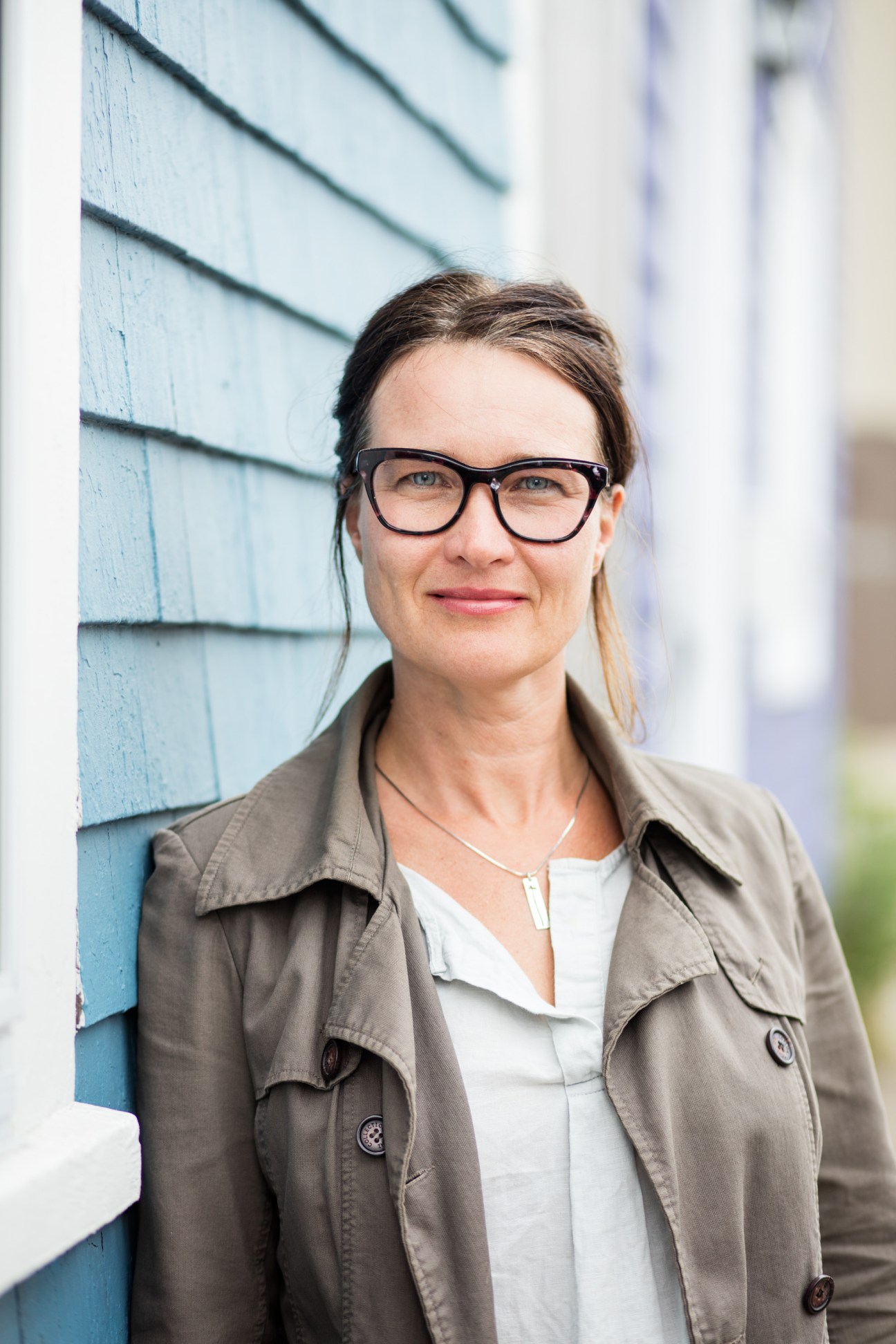 Patricia Cuttell stands outdoors wearing glasses and a beige jacket against blue shingled siding