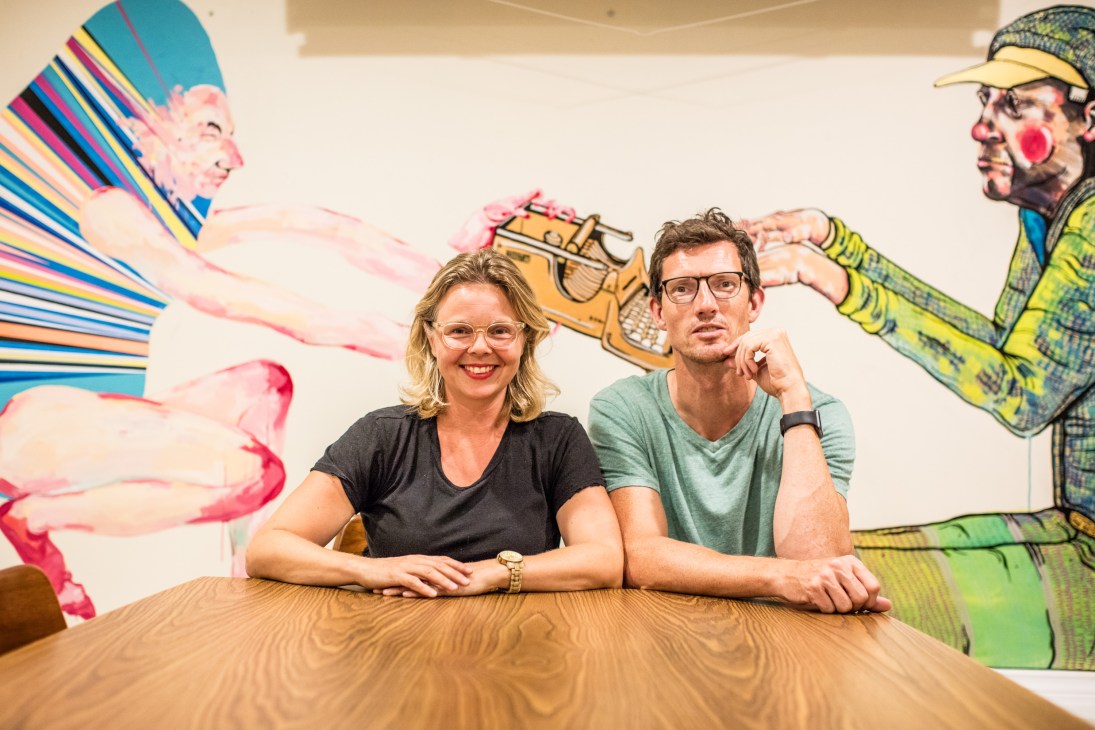 Two people sitting at a wooden table in front of a colorful mural with stylized figures