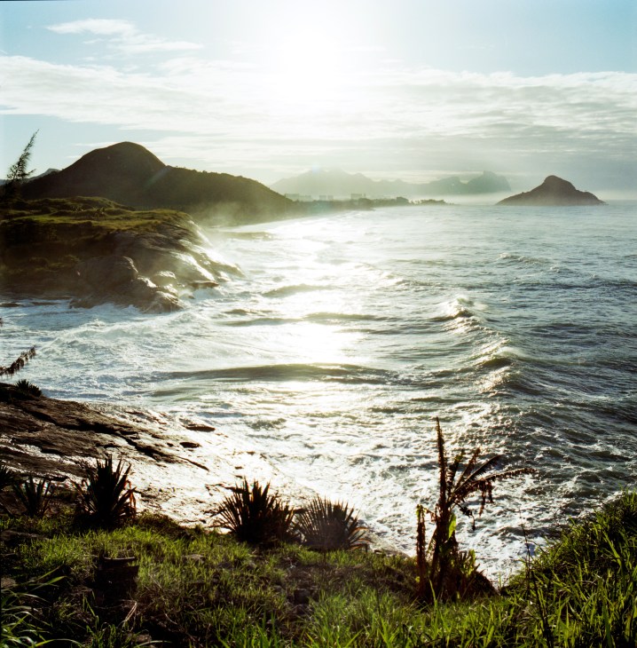 Macumba beach with rolling waves, rocky outcrops, tropical vegetation, and mountains visible across the water.