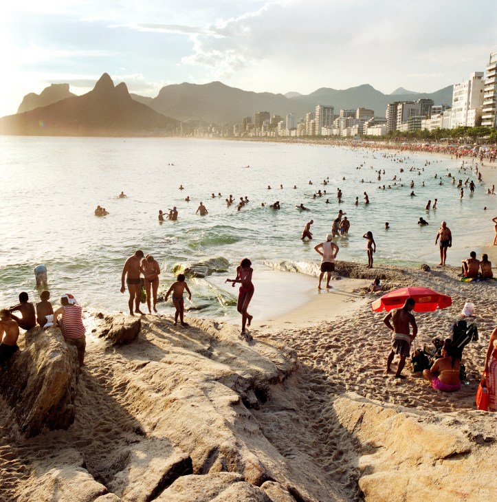 Crowded Ipanema beach with swimmers and sunbathers, mountains visible across the bay.