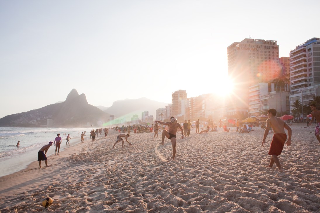 People playing football on a sandy beach at sunset with Rio's mountains and high-rise buildings in the background.