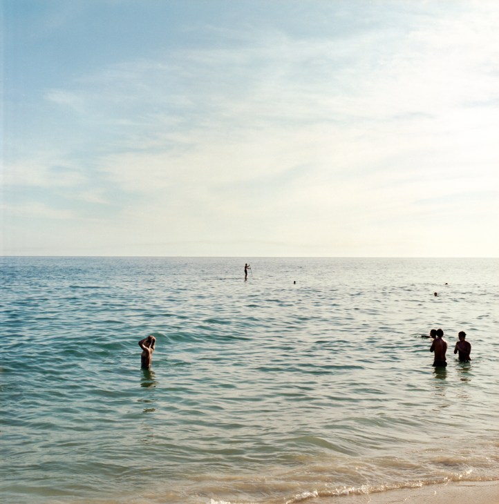 People swimming and wading in calm turquoise waters off a Rio de Janeiro beach under a clear sky.