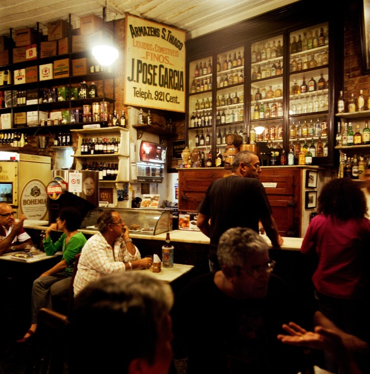Interior of Bar do Gomes in Santa Teresa with patrons at tables and vintage liquor bottles displayed on shelves behind the bar.