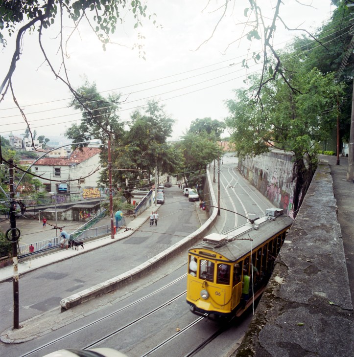 Yellow tram car on tracks climbing a steep street lined with trees and colonial buildings in Santa Teresa, Rio de Janeiro.