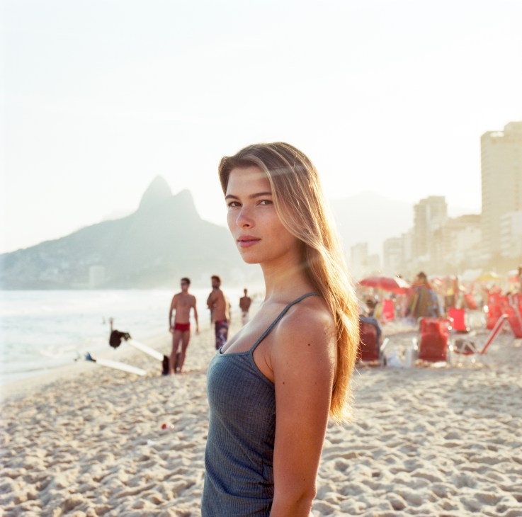 Young woman in blue swimsuit standing on Ipanema beach with mountain in background.