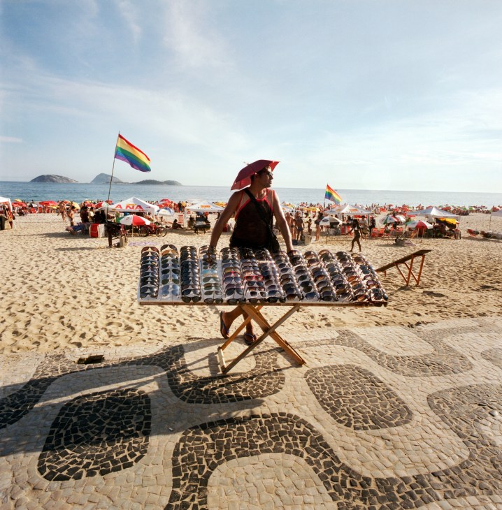 Vendor selling sunglasses on Ipanema beach with rainbow flags and beachgoers in background.