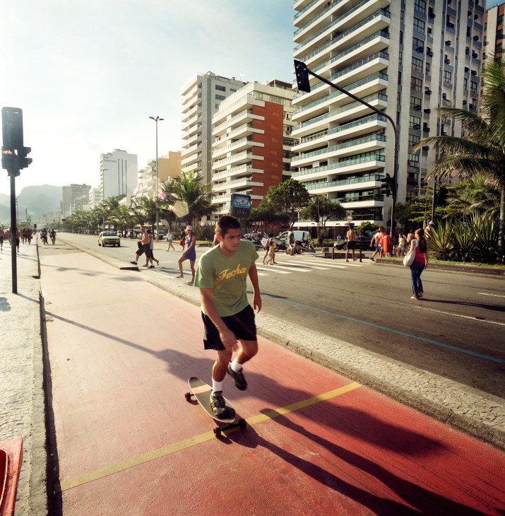 A skateboarder rides along the red-painted Ipanema promenade with beachfront apartment buildings and pedestrians behind.