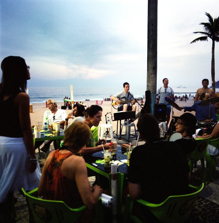 Musicians perform on a beach as beachgoers gather around green tables to listen and socialize.