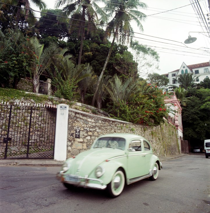 A mint-green Volkswagen Beetle drives past colonial architecture and lush vegetation on a Rio street.