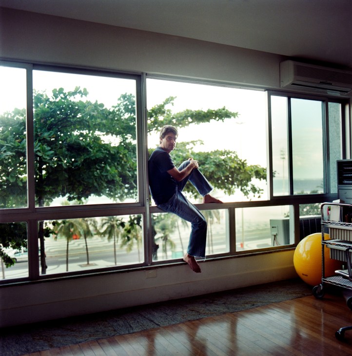 Director Andrucha Waddington sits on a windowsill in his apartment overlooking Leme beach in Rio de Janeiro.