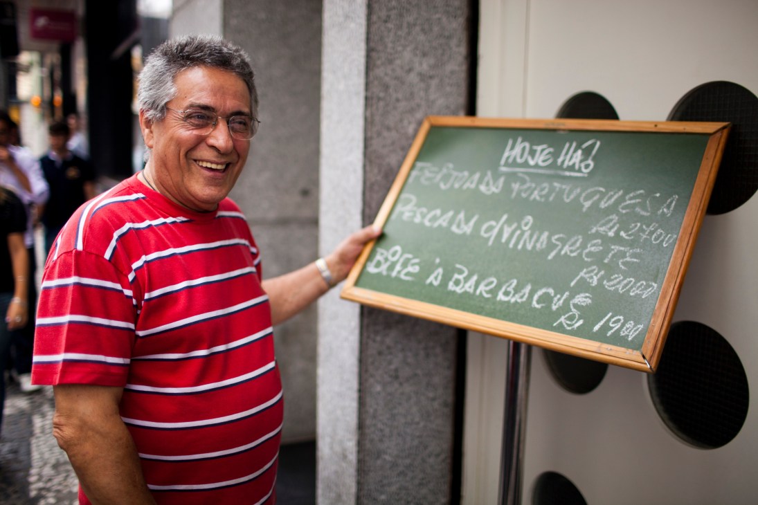 José Paiva, owner of Cais da Ribeira Portuguese restaurant, smiles while holding a chalkboard menu on a city street.