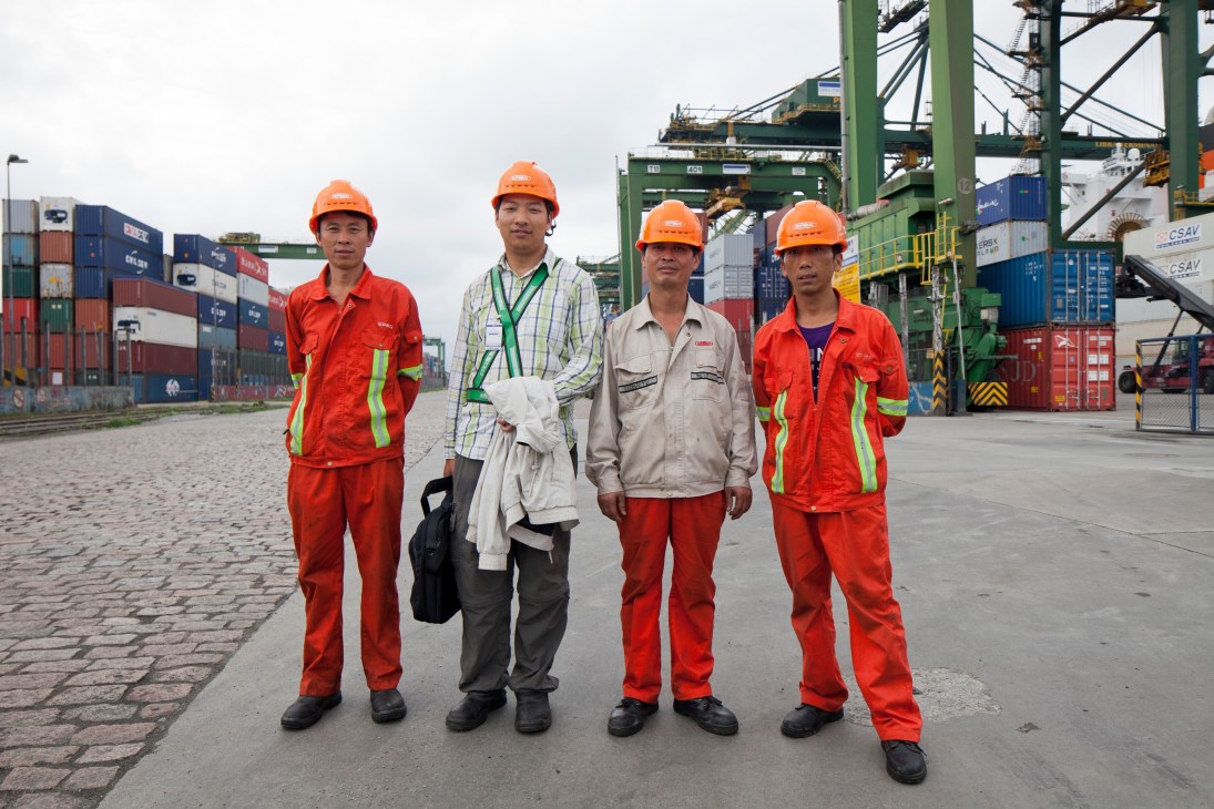 Four workers in safety gear stand at Santos port with shipping containers and cranes in the background.