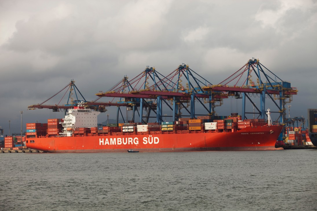 Red Hamburg Süd container ship docked at Santos port with blue cargo cranes and stacked containers.