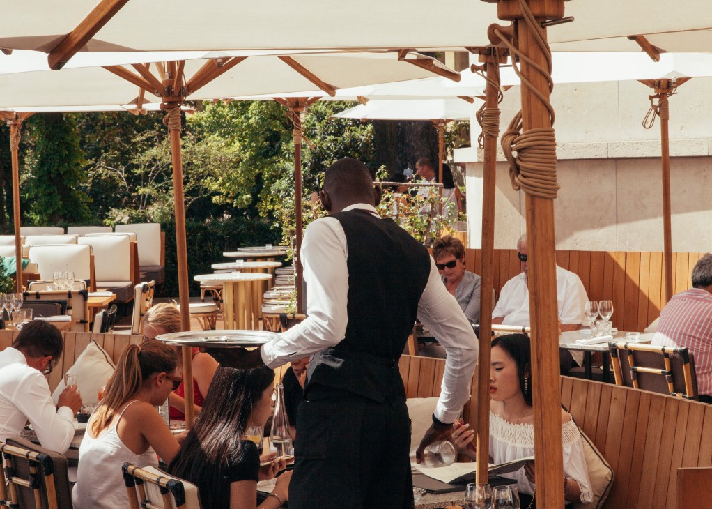 Waiter serving dishes on a wooden pergola-covered terrace at Café de l'Homme restaurant.