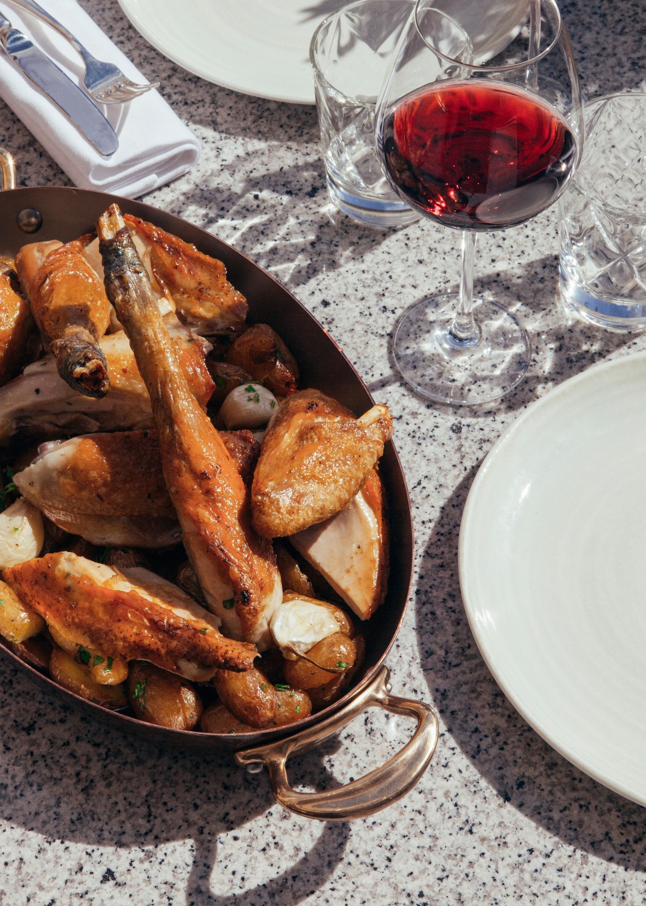 Roasted Bresse chicken with grenaille potatoes and garlic served in a copper pan with red wine and place settings.