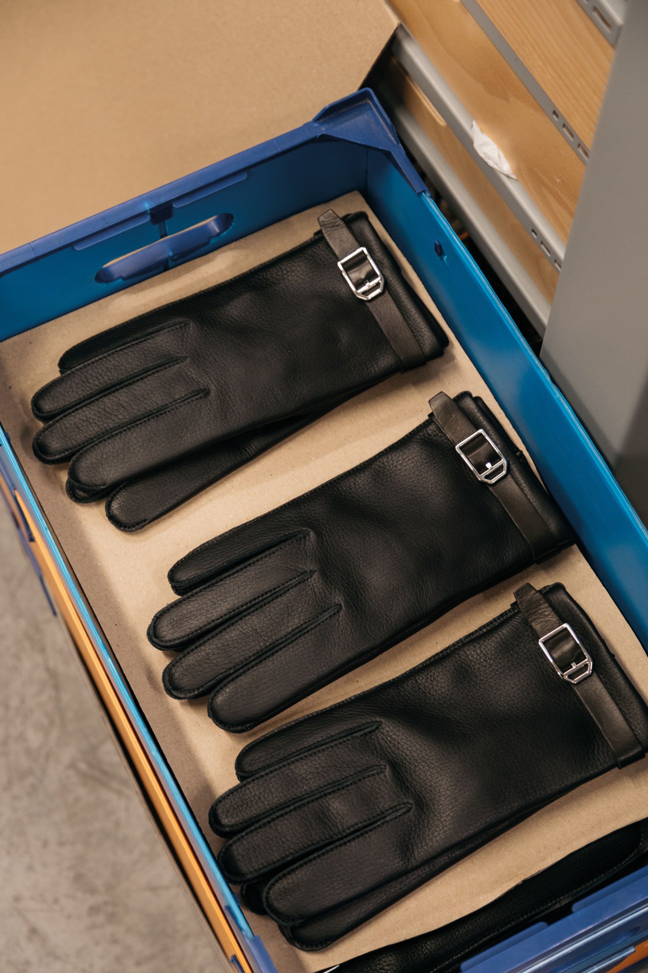 Three pairs of black leather gloves with metal buckle details displayed in a blue Hermès box with beige linen compartments.