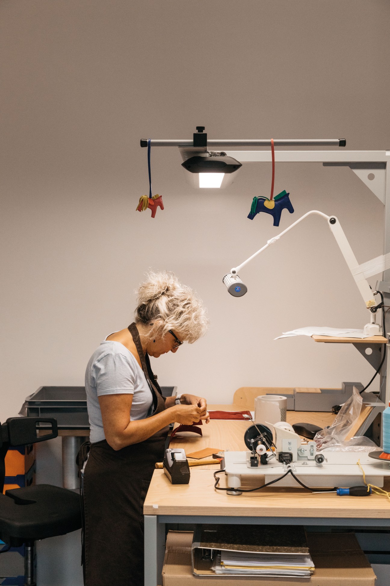 Artisan working at a wooden workbench in a modern workshop with pendant lighting and Hermès horse charms.