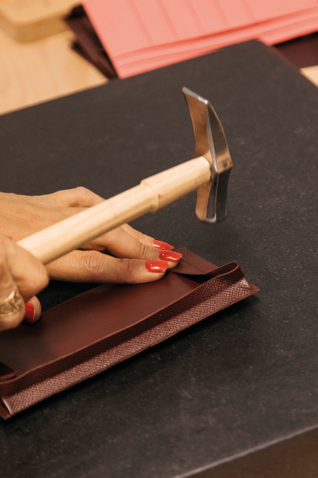 Hands holding a hammer and iron hand tool over brown leather on a black workbench.
