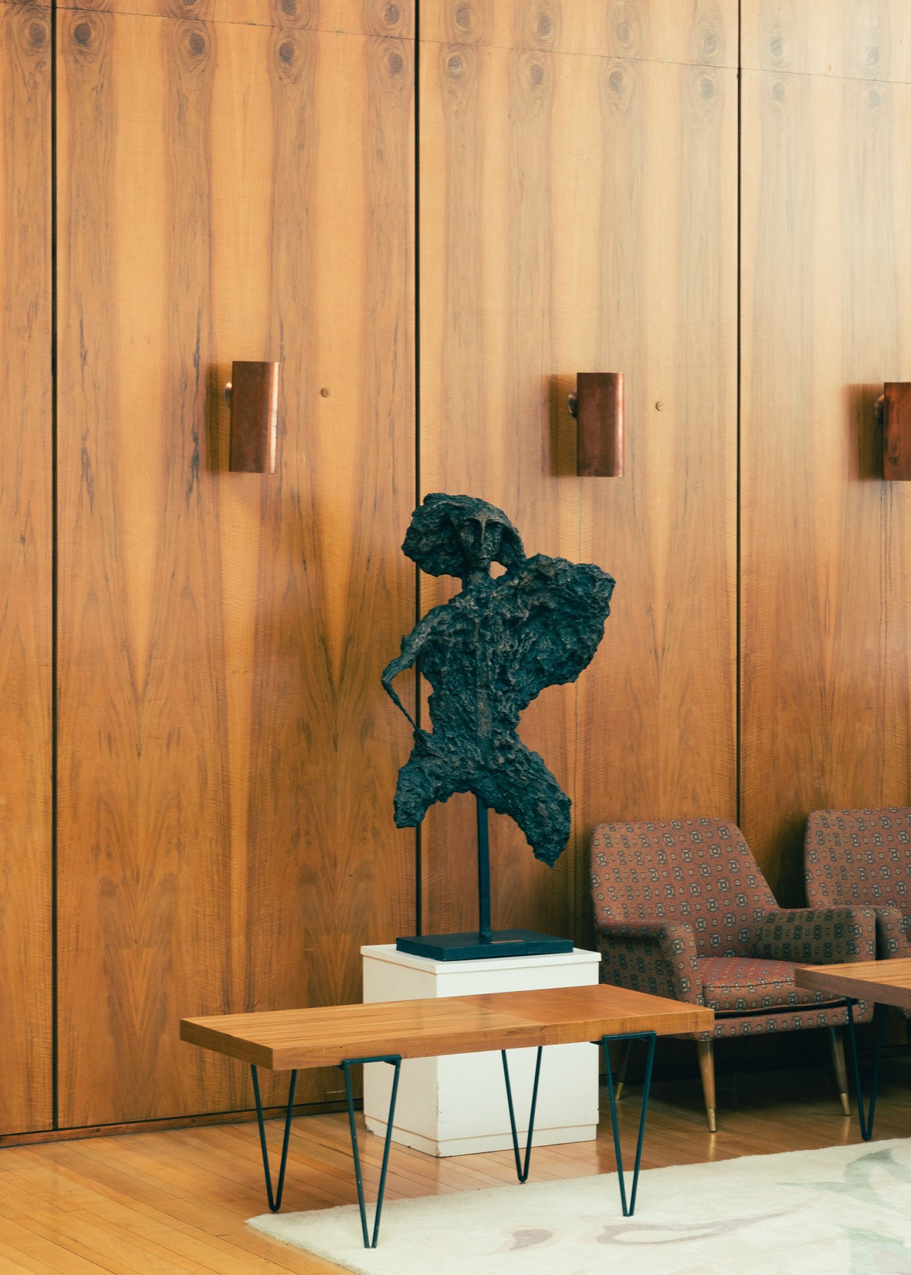 Dark teal sculpture displayed on white pedestal in modernist interior with wood paneling and mid-century furniture.