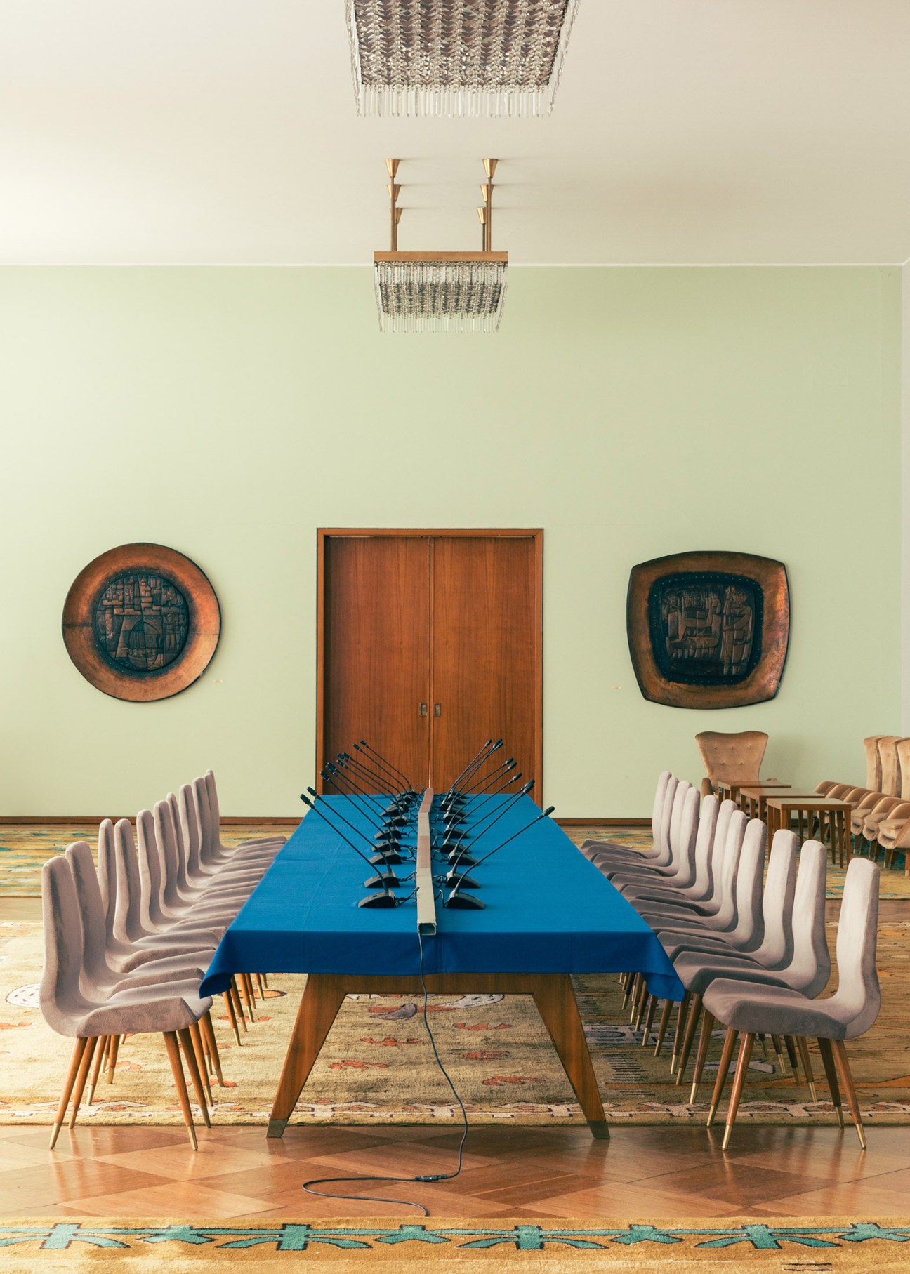 Mid-century modern conference table with blue top and wooden chairs in pale green room with copper relief plaques.