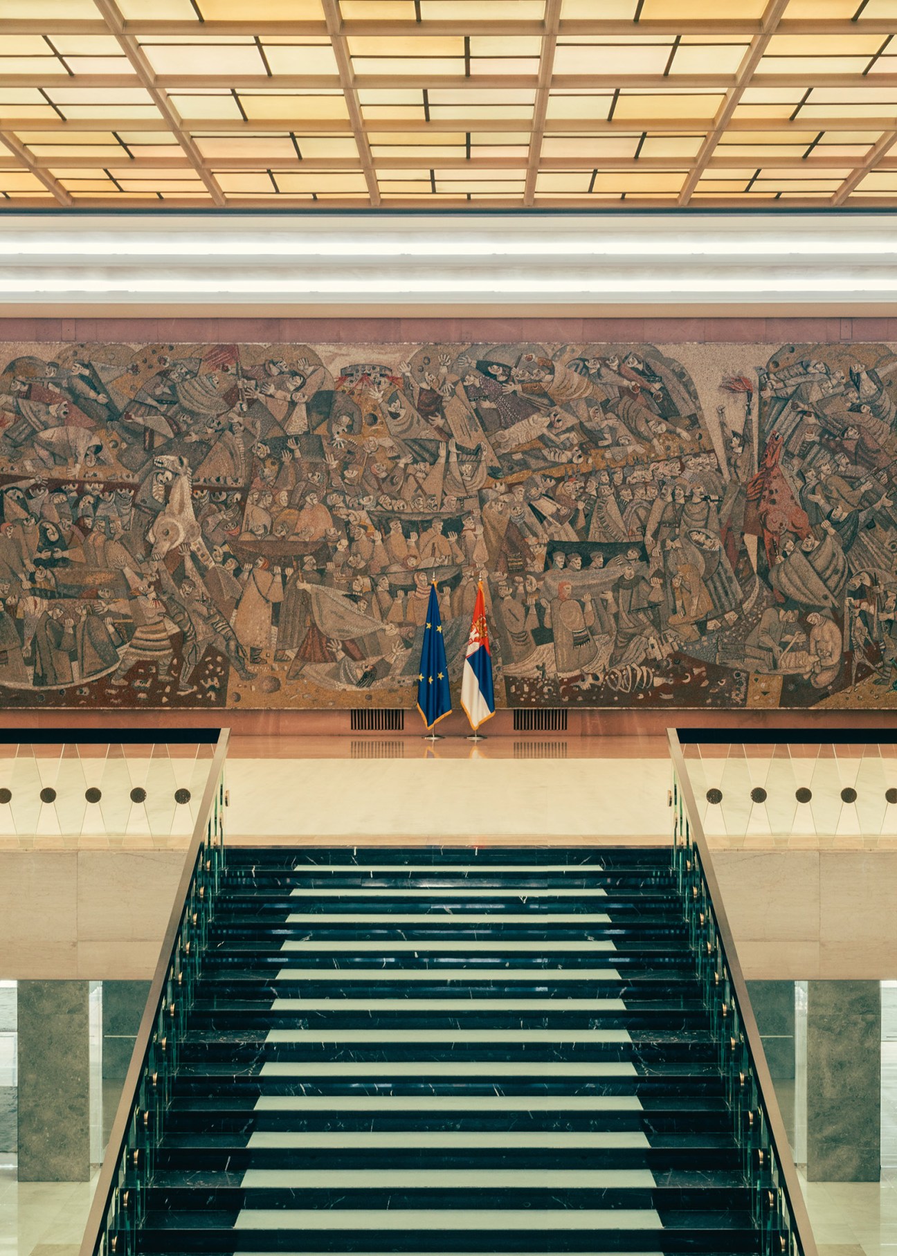 A grand modernist staircase with teal steps leads upward toward a large expressionist mural featuring figures and animals, flanked by EU and Serbian flags.