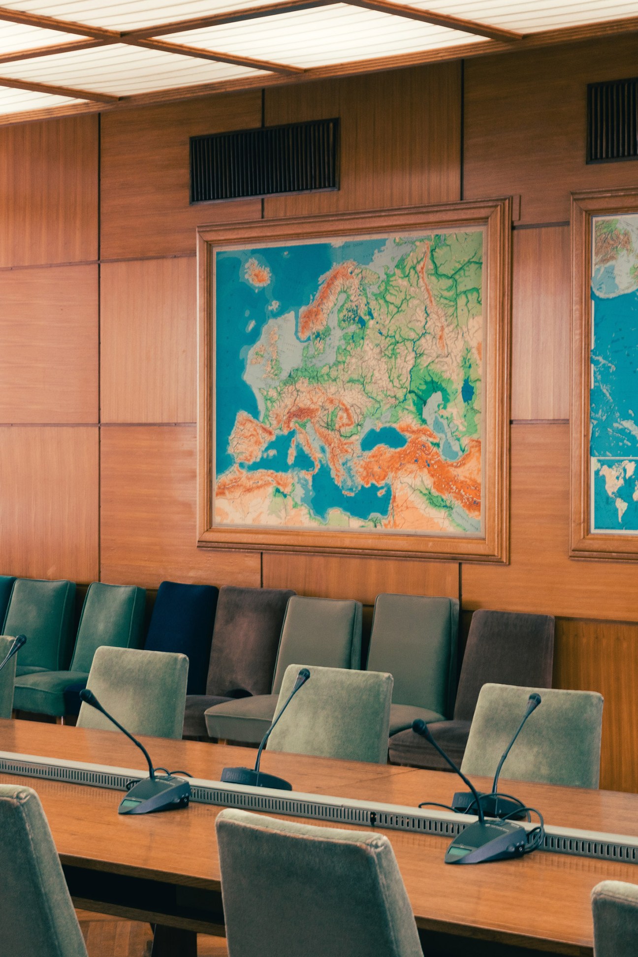 Mid-century modern conference room with wood paneling, framed relief map of Europe, and upholstered chairs at wooden table.