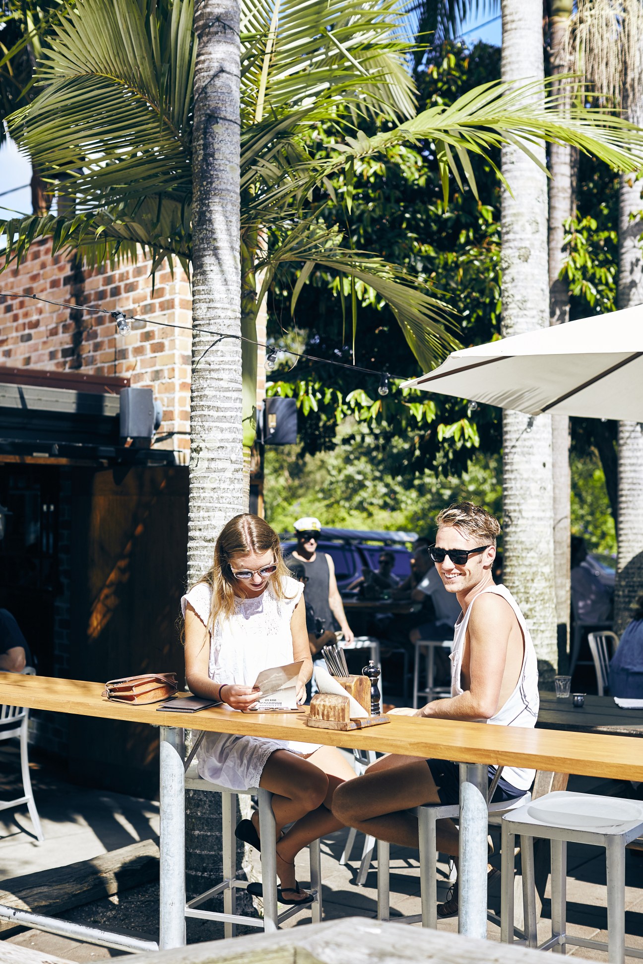 Two people dining outdoors at a wooden table under palm trees and a white shade awning at The Roadhouse.