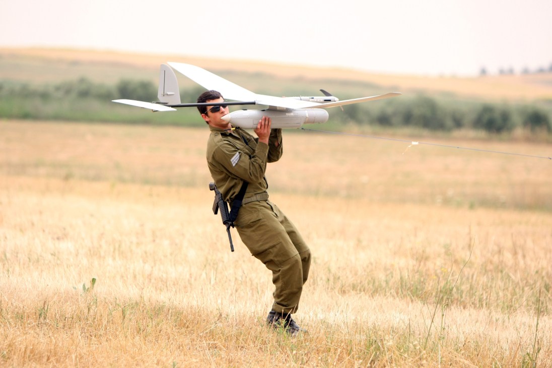 Israeli soldier in olive uniform launching a white surveillance drone in an open field.