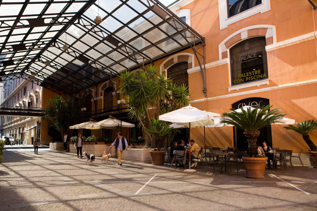 Orange building courtyard with glass roof structure, white umbrellas, and people dining outdoors in Rome.