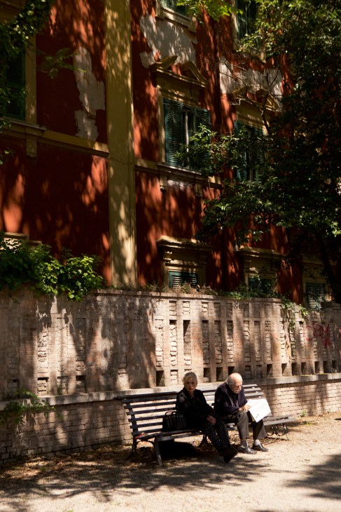 Two elderly men sit on a bench in dappled shade beneath a terracotta building covered in climbing vines.