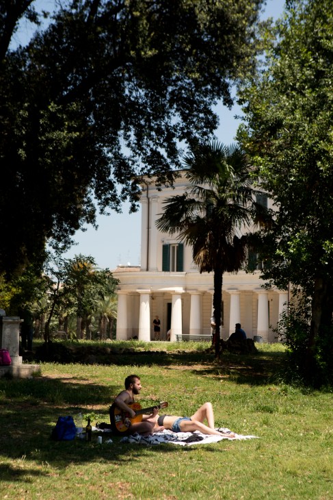 Man playing guitar on grass in Villa Torlonia park with neoclassical building and palm trees in background