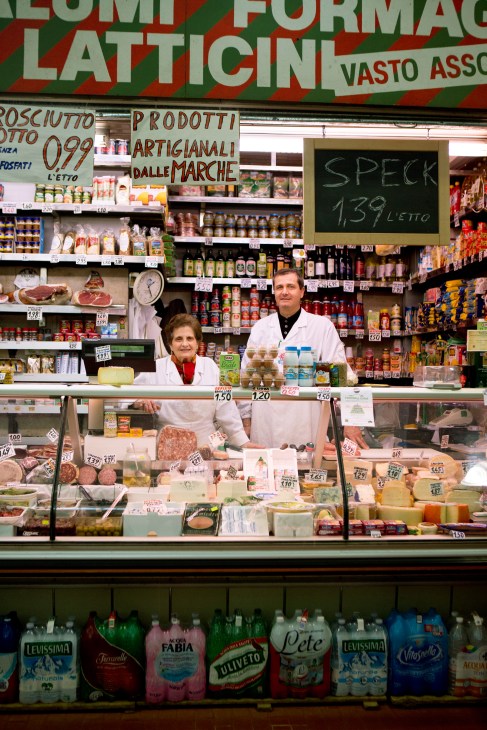 Two butchers in white coats stand behind a deli counter displaying cured meats and cheeses in Rome's Trieste market.