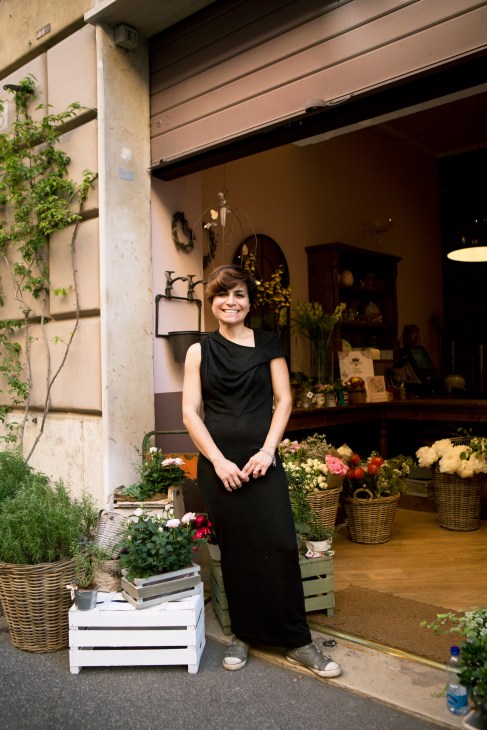 Woman in black sleeveless dress standing outside a flower shop storefront with potted flowers.