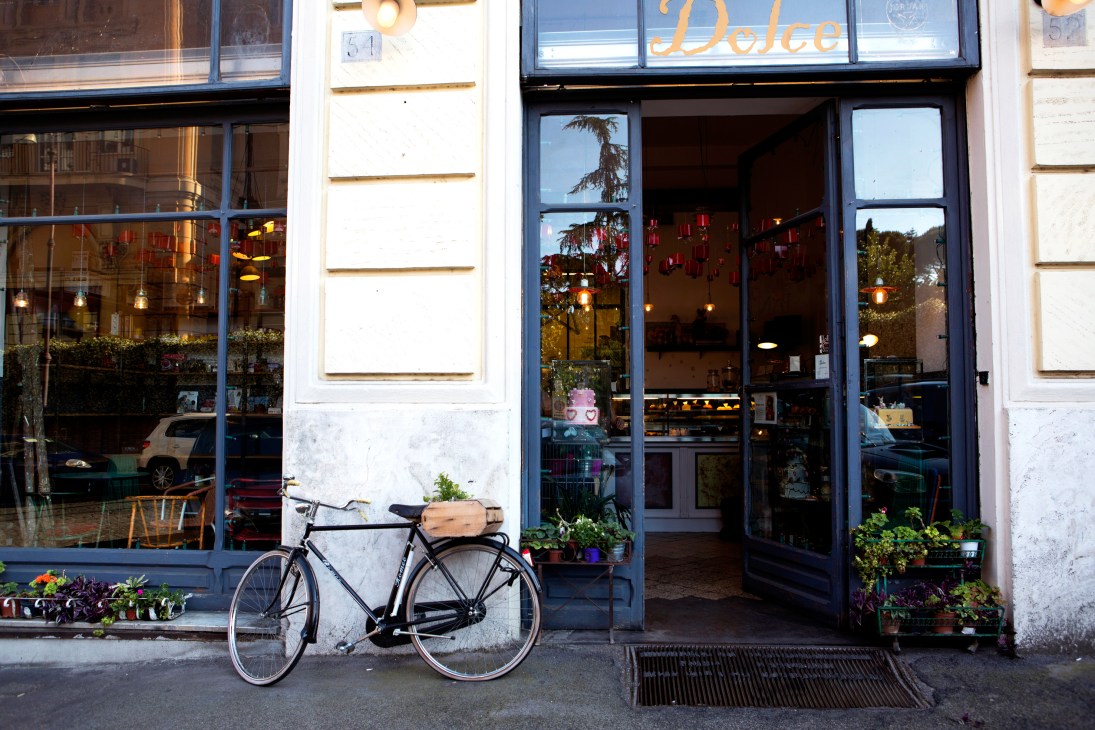 Bicycle parked outside Dolce pastry shop with blue storefront and hanging lights.