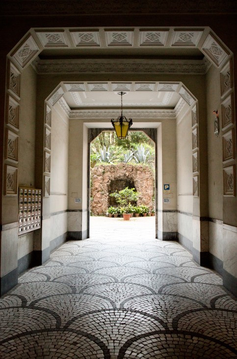 Ornate neoclassical archway with decorative ceiling panels and yellow pendant lamp opening to a courtyard garden.