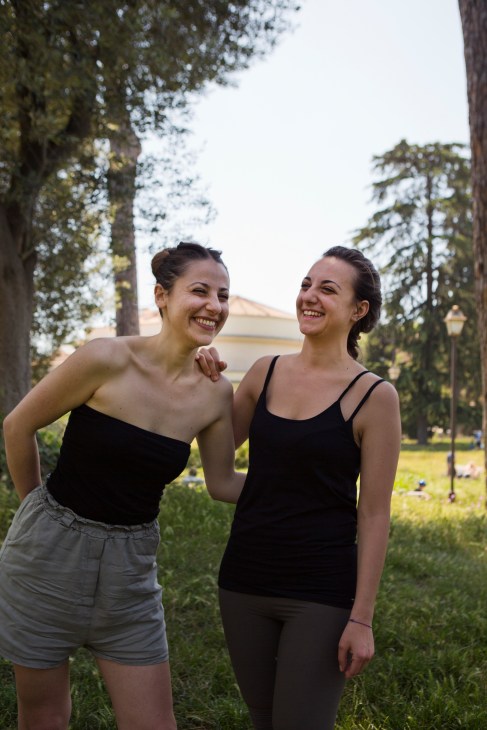 Two young women smiling in Villa Torlonia park with tall pine trees and green grass behind them.
