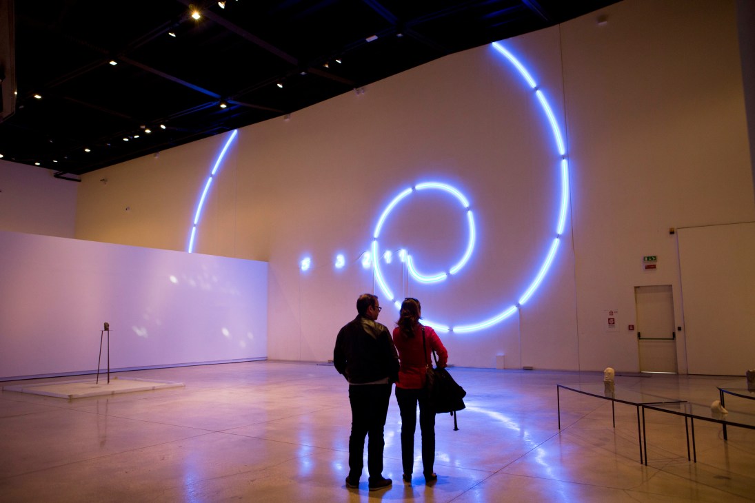 Two visitors stand in a modern art gallery with blue neon circular light installations on white walls.