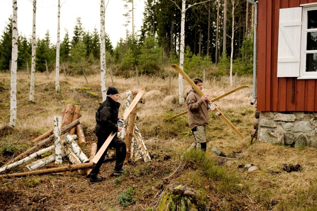 Two men carry wooden skis through a forest of birch and pine trees near a red wooden building.