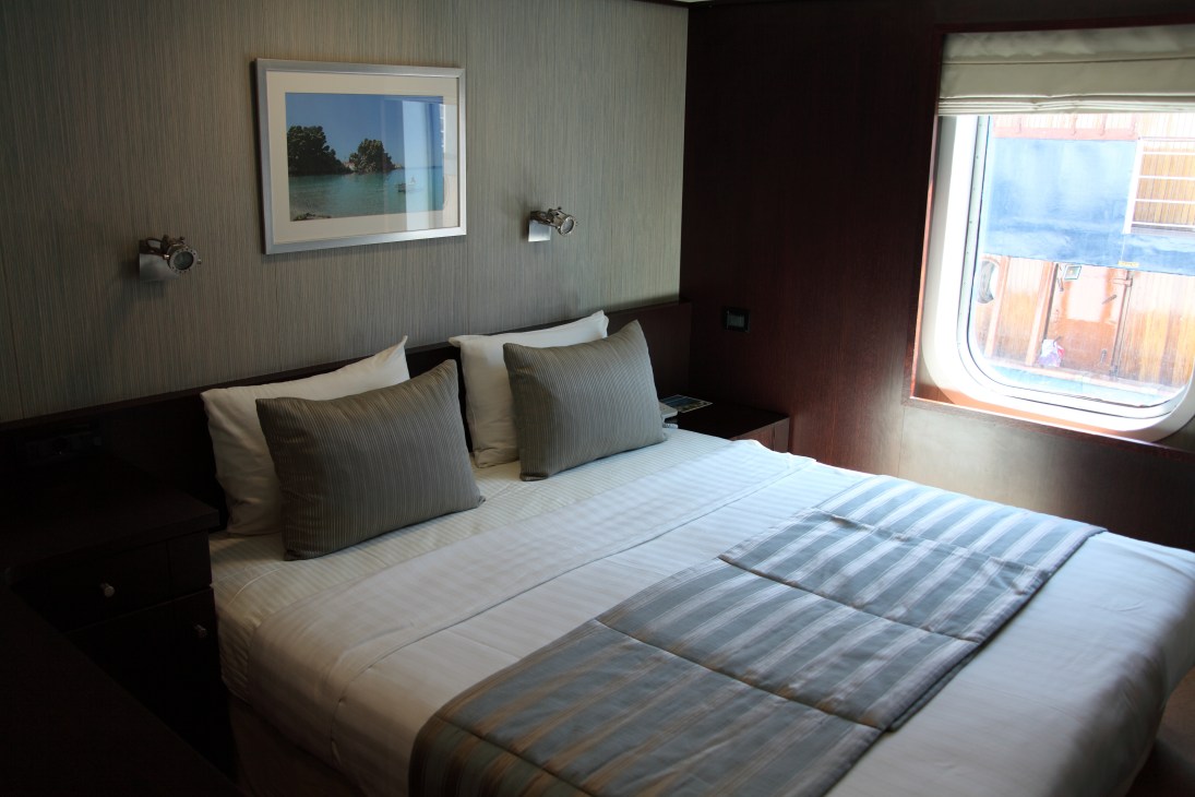 A modern cruise ship cabin with a double bed, neutral décor, and a porthole window overlooking the sea.