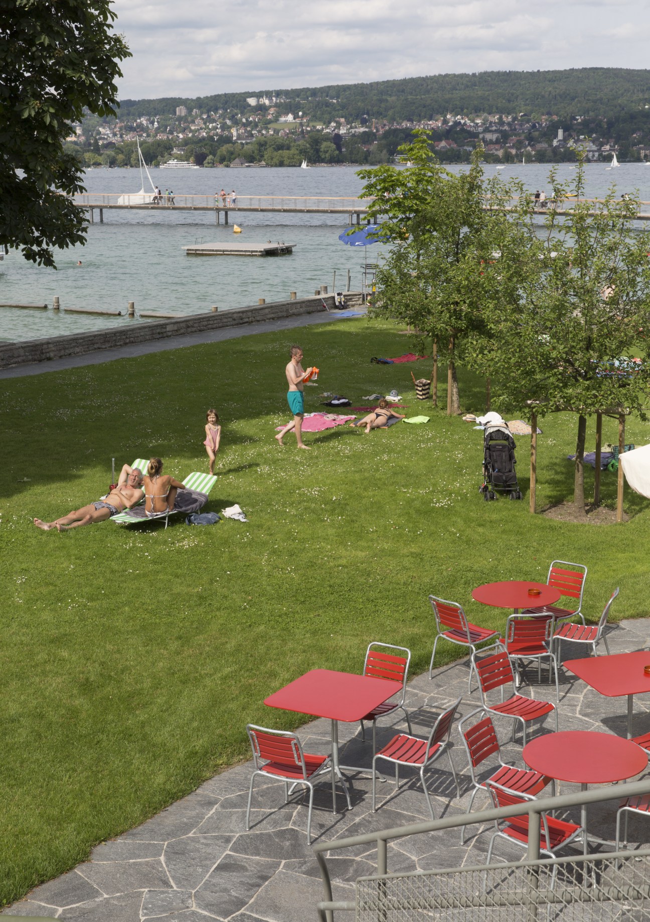 Visitors relax on the grass at Strandbad Wollishofen with Lake Zürich and surrounding hills in background.